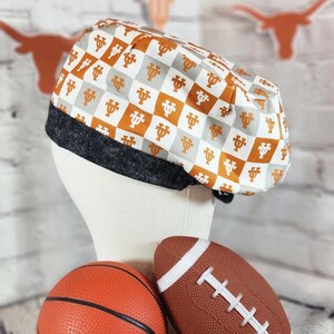 May include: A patterned scrub cap with a black brim, featuring a repeating design in orange and gray on a white background. A basketball and a football are in the foreground.