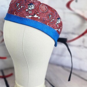May include: A scrub cap featuring a maroon fabric with a repeating pattern of the SC State University bulldog mascot and circular logos. The cap has a solid blue fabric band along the bottom edge and includes black ties extending from the back side.