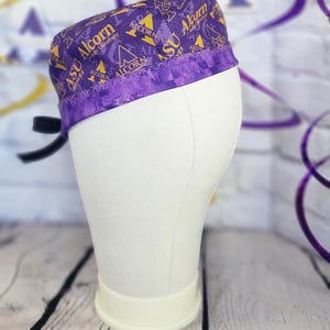 May include: A purple scrub cap featuring a repeating pattern of the gold Alcorn State University logo and text. The cap has a wide, solid purple band along the bottom edge with a decorative zigzag stitch and black ties extending from the back.
