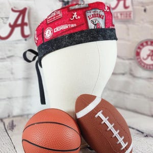 May include: A red and black patterned scrub cap with the text "Crimson Tide" and "Alabama" logos. A basketball and a football are in front of the cap. The cap is tied in the back with black ties.