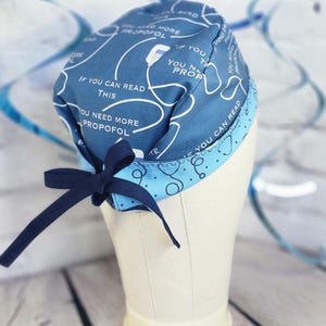 May include: A blue scrub cap featuring a repeating pattern of medical IV bags and the text IF YOU CAN READ THIS YOU NEED MORE PROPOFOL. The cap has a lighter blue trim with a stethoscope print and a dark blue fabric tie at the back for adjustment.