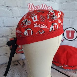 May include: A red and white patterned scrub cap featuring the Utah Utes logo and helmet graphics. The cap has a red brim and black ties. A matching red cap is also visible.
