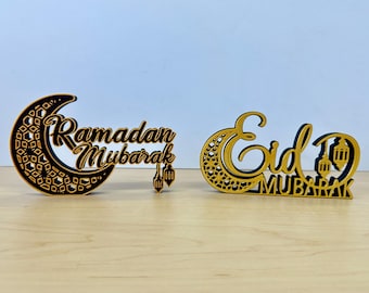 May include: Two wooden signs with crescent moons and Arabic calligraphy. The first sign says "Ramadan Mubarak" and the second sign says "Eid Mubarak".