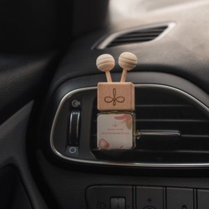 Könnte beinhalten: Ein hölzerner Autolufterfrischer mit zwei Holzperlen oben. Der Lufterfrischer befindet sich in einer Lüftung eines Autos und hat ein Etikett mit der Aufschrift "JUNAA HOME CREAM STRAWBERRY & RAIN".