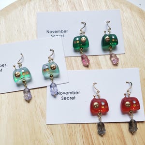May include: A selection of earrings in various colours, including green, blue, and red, with a controller design and gold detailing. Each earring has a dangling charm. The text "November Secret" is printed on the cards.