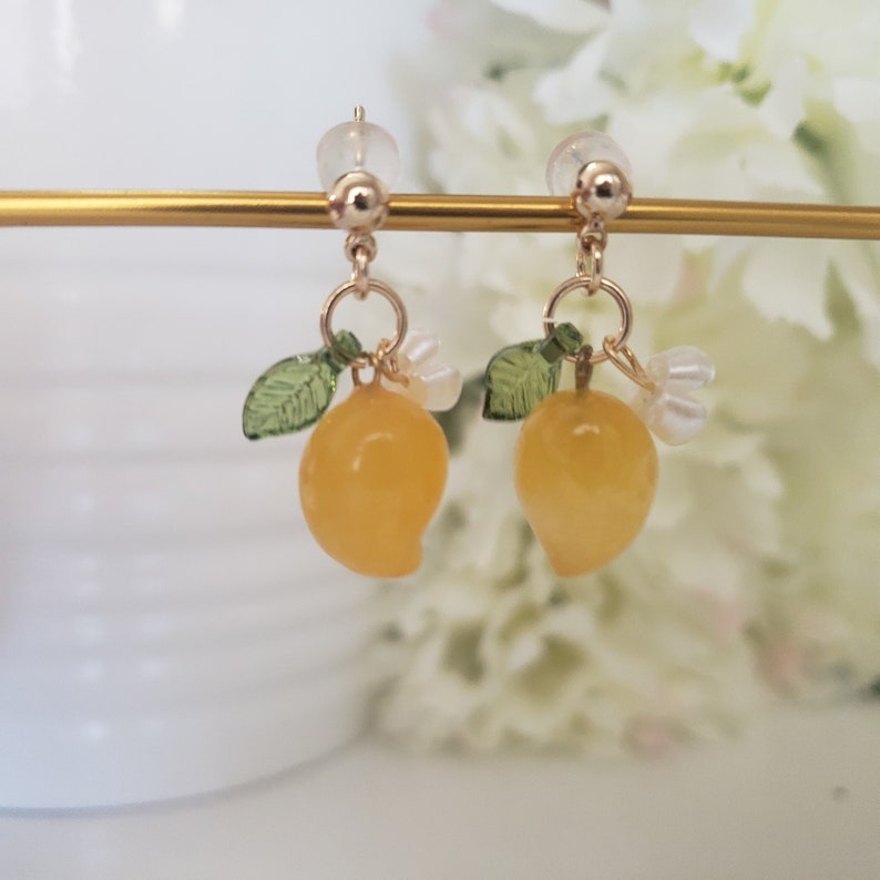 May include: A pair of gold earrings with yellow lemon charms, green leaves, and white pearl accents.