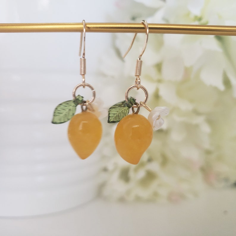 May include: A pair of gold earrings with a yellow lemon charm, green leaves, and white flower accents.