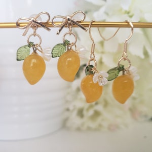 May include: Gold earrings with a lemon charm, green leaves, and white flower accents. The earrings are on a gold stand.