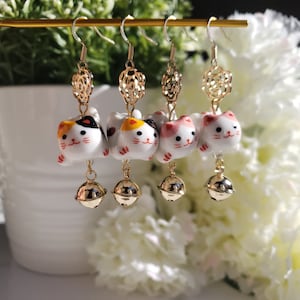 Pode incluir: Quatro pares de brincos de gato de ouro com um pingente de sino. Cada amuleto de gato é branco com detalhes pretos, amarelos e rosados.