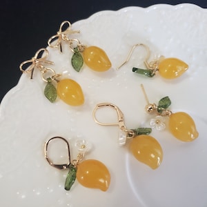 May include: Five pairs of lemon-shaped earrings with gold-tone findings. Each earring features a yellow lemon charm with a green leaf and a gold-tone accent. Two pairs have a gold-tone bow at the top.