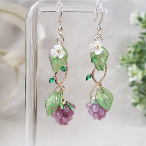 May include: Gold-tone dangle earrings featuring a grape cluster design. Each earring has a cluster of purple grapes, green leaves, white flowers, and green accents. The earrings are suspended from a gold-colored hook.
