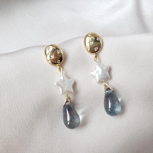 May include: Elegant gold-tone earrings. Each earring has a gold oval stud with star details, a white star charm, and a blue-grey teardrop stone. The earrings hang down, creating a delicate and elegant appearance. The earrings are suitable for special occasions.