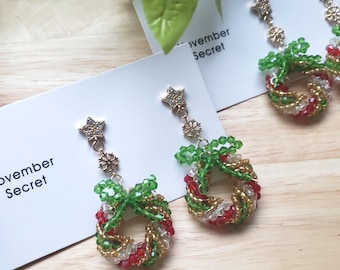 May include: Dangle earrings featuring a beaded wreath design in red, green, and gold. Each earring has a gold-tone star stud with a small snowflake charm attached to a chain. The white display card features the text November Secret.