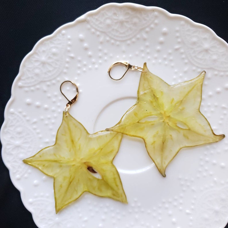 May include: A pair of starfruit earrings. Each earring is a translucent, star-shaped slice of yellow fruit with gold-colored ear wires. The earrings are displayed on a white, patterned plate.