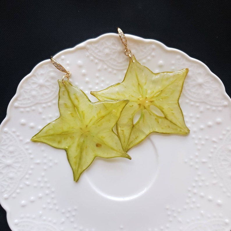 May include: A pair of starfruit earrings. Each earring is a translucent, star-shaped slice of fruit, preserved in resin. The earrings hang from gold-colored hooks with small, sparkling accents. The earrings rest on a white, patterned plate.