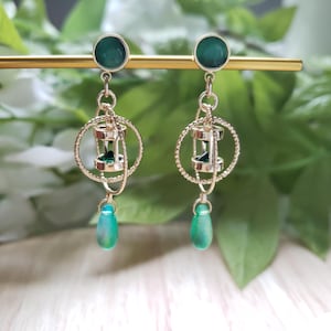 May include: Gold-toned earrings with a green circular stud, a circular frame, and a miniature hourglass design. The earrings have a teardrop-shaped green bead dangling from the bottom. The background is blurred with green foliage.