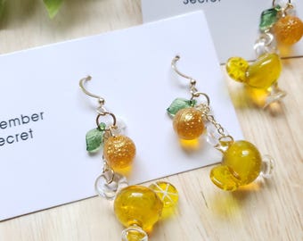 May include: A pair of handmade earrings featuring a cocktail-themed design. The earrings have gold-colored hooks, with clear and yellow glass beads, and small green leaf accents. The earrings are on a white card with the text "November Secret".