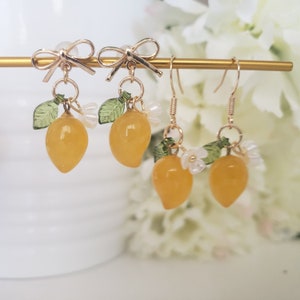 May include: A pair of gold lemon earrings with white flower accents and green leaves. The earrings are on a gold stand.