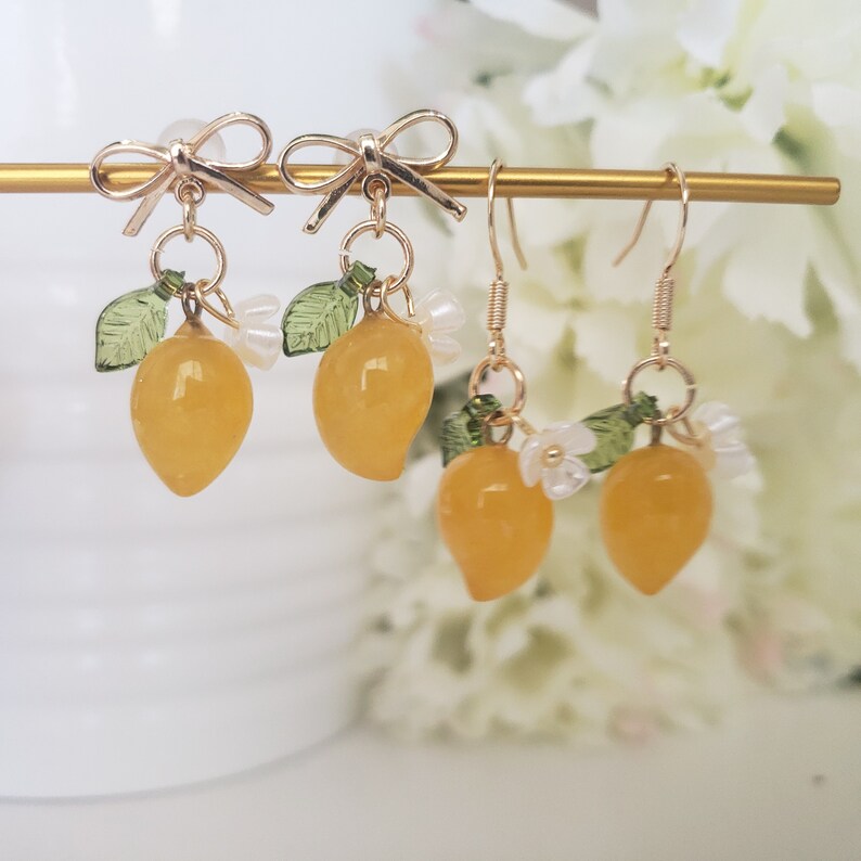 May include: A pair of gold lemon earrings with white flower accents and green leaves. The earrings are on a gold stand.