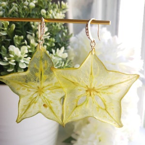 May include: A pair of gold-toned earrings featuring star-shaped pendants. The pendants are a translucent yellow, resembling star fruit slices. The earring hooks are adorned with small, sparkling crystals, adding a touch of elegance to the unique design.
