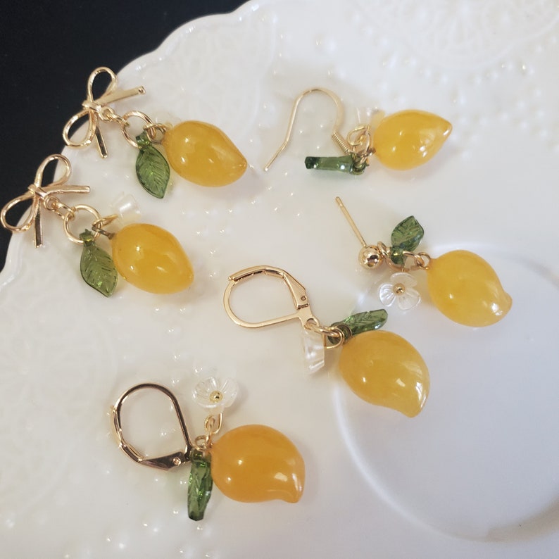 May include: A set of five pairs of lemon-shaped earrings with gold-tone findings. Each earring features a yellow lemon charm with green leaves and a small white flower.