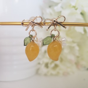 May include: Gold earrings with a lemon charm, green leaves, and white flowers. The earrings have a bow at the top.