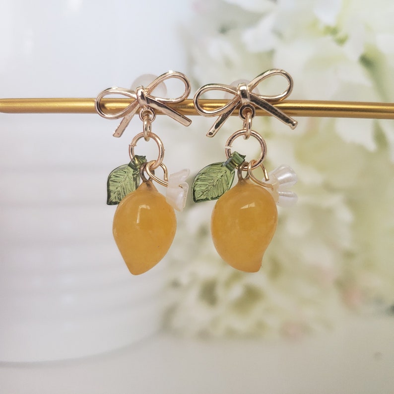 May include: Gold earrings with a lemon charm, green leaves, and white flowers. The earrings have a bow at the top.
