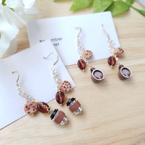 May include: Two pairs of dangle earrings featuring miniature charms of chocolate chip cookies, dark brown coffee beans, and coffee cups. The earrings are suspended from silver-toned hooks with small clear beads on the chains, displayed on white cards.