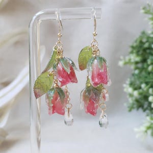 May include: Dangle earrings featuring pink and clear resin rose buds with green leaves and gold-colored metal accents. Each earring has three rose buds and a clear, teardrop-shaped charm. The earrings are displayed on a clear stand.