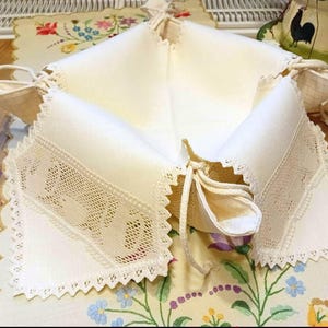 Può includere: Un cestino in tessuto piegato color crema con bordi smerlati e dettagli in pizzo. Il cestino è legato con nastri color crema. Lo sfondo include una tovaglia a motivi floreali e una tazza da tè decorativa a tema gallo.