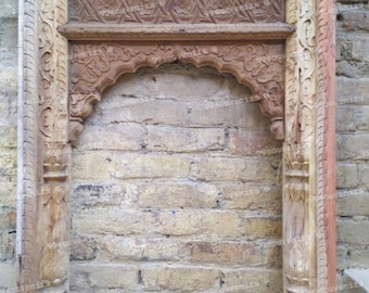 May include: A carved wooden frame with intricate details, featuring a rounded archway. The frame is made of light and dark wood, creating a contrasting effect. The frame is set against a brick wall.