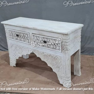 May include: White wooden console table with two drawers and intricate carved details. The table has a distressed white finish and a unique arched base.
