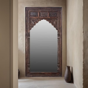 May include: A large, ornate wooden mirror with intricate carvings and an arched top. The mirror is set against a light brown wall.