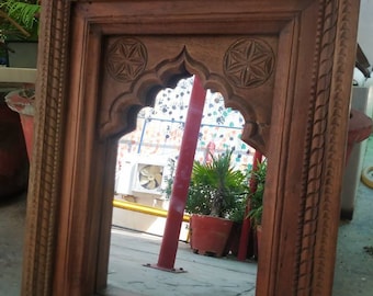 May include: A wooden mirror with a carved frame and an arched top. The frame is intricately carved with floral and geometric designs. The mirror is reflecting a view of a courtyard with plants and a red metal pole.