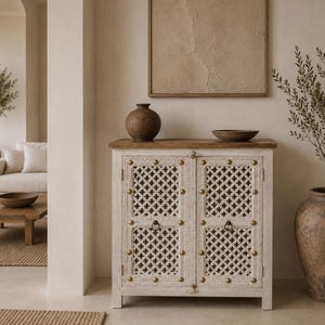 Puede incluir: Un mueble rectangular de madera con dos puertas que presentan un enrejado blanco intrincado y tachuelas de metal en tono latón. El mueble tiene una superficie superior de madera natural lisa y pequeños tiradores de anillo de metal negro.