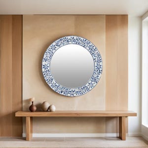 May include: A round mirror with a blue and white floral design. The mirror is hanging on a light brown wall above a wooden bench.