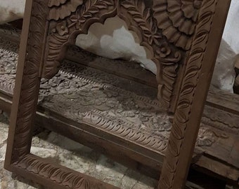 May include: A dark brown wooden frame with intricate carvings of leaves and flowers. The frame has an arched opening at the top and is designed to hold a picture or mirror.