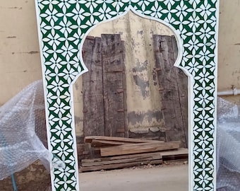 May include: Rectangular mirror with a white frame and a green and white floral pattern border. The mirror has an arched top and reflects a view of wooden planks and a wall.