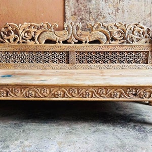May include: A wooden bench with intricate carvings of peacocks and floral designs. The bench has a slatted back and a solid seat. The wood is a light brown color.
