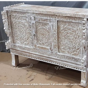 May include: Whitewashed wooden cabinet with intricate carvings and three doors. The cabinet has a distressed finish and sits on four legs.