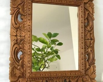 May include: A rectangular wooden mirror with an intricately carved frame. The frame is a warm brown colour and features detailed floral and scrollwork designs. The mirror reflects a green plant. A decorative home decor item.