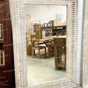 Könnte beinhalten: Ein großer, weißer, verzierter Holzspiegel mit einem rechteckigen Rahmen. Der Rahmen ist mit komplizierten Details geschnitzt und hat eine abgenutzte Oberfläche. Der Spiegel spiegelt einen Raum mit Möbeln wider.