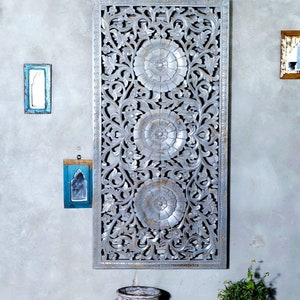 May include: A silver-painted wooden wall decor with intricate floral carvings and three circular designs. The decor is hanging on a gray wall with a white vase and a small plant in a brown pot.