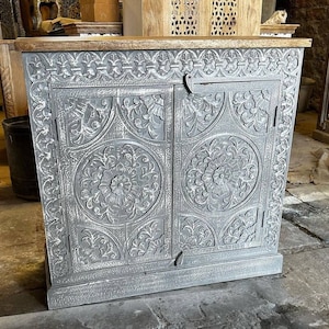 Puede incluir: Un armario de madera gris con dos puertas. Las puertas están talladas con intrincados motivos florales y tienen un acabado blanco desgastado. El armario tiene una tapa de madera y una base.