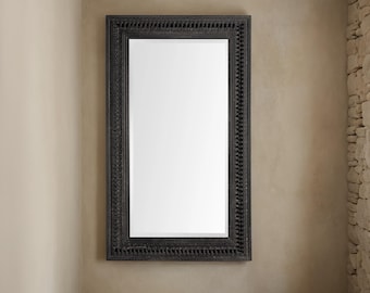 May include: A black framed mirror with an ornate design. The mirror is hanging on a beige wall.