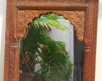 May include: A wooden mirror with intricate carvings around the frame. The mirror is arched at the top and reflects a potted plant with green leaves.