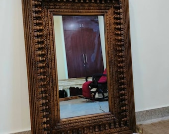 Puede incluir: Un espejo de madera grande y tallado intrincadamente con un acabado marrón. El espejo tiene una forma rectangular y un marco detallado con un patrón repetitivo. El espejo está apoyado contra una pared.