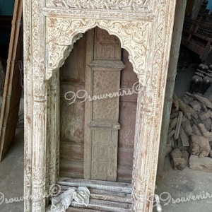 Puede incluir: Un arco de madera blanquecina envejecida con intrincadas tallas. El marco presenta patrones florales y geométricos, con una abertura arqueada y un aspecto desgastado. La pieza tiene una estética rústica y vintage.