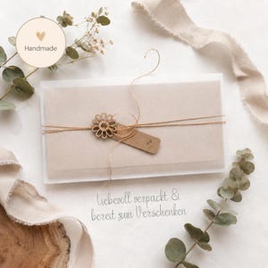 Pu&ograve; includere: Una busta rettangolare in carta velina traslucida, avvolta da uno spago sottile e fissata con un piccolo ciondolo in legno a forma di fiore e un'etichetta rettangolare vuota. Il testo "Handmade" appare in un cerchio, con "Liebevoll verpackt & bereit zum Verschenken" stampato sotto.
