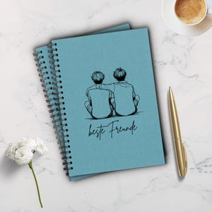 Könnte beinhalten: Ein blaues Spiralnotizbuch mit einer schwarz-weißen Illustration von zwei Personen, die sich umarmen. Unter der Illustration steht der Text "beste Freunde".